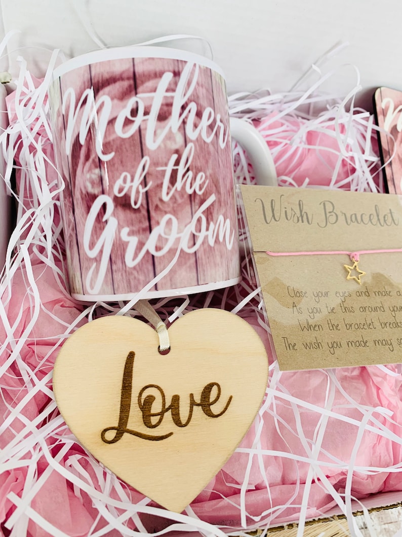 May include: A gift box filled with pink tissue paper and a white mug with the text "Mother of the Groom" printed on it. The box also contains a wooden heart with the word "Love" engraved on it and a wish bracelet with a star charm.
