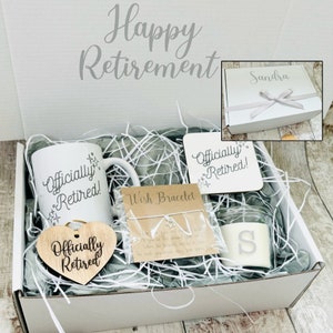 May include: A white gift box filled with retirement themed gifts. The box contains a white mug with the text "Officially Retired!" a wooden heart with the same text, a small wooden sign with the same text, a small candle with the letter "S" on it, and a wish bracelet.