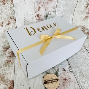 May include: A white gift box with a gold ribbon and the name "Denice" printed on the top. A small wooden circle with the text "Countdown & Celebrate" is in the foreground.