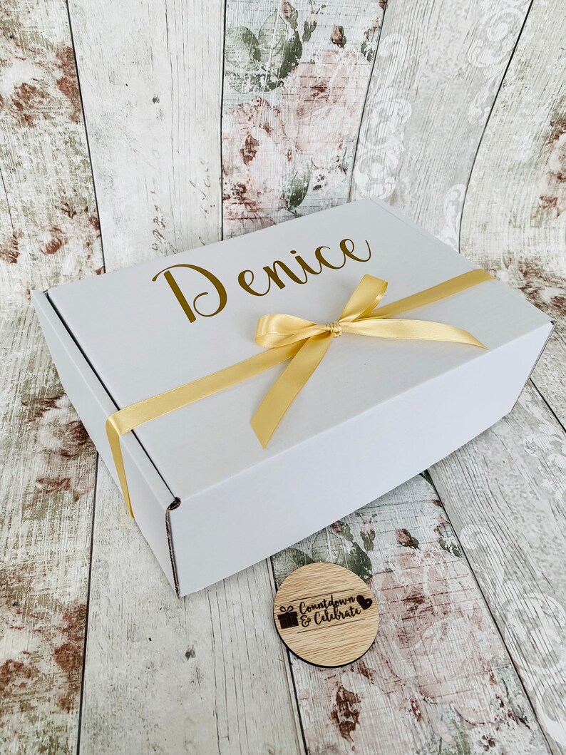 May include: A white gift box with a gold ribbon and the name "Denice" printed on the top. A small wooden circle with the text "Countdown & Celebrate" is in the foreground.