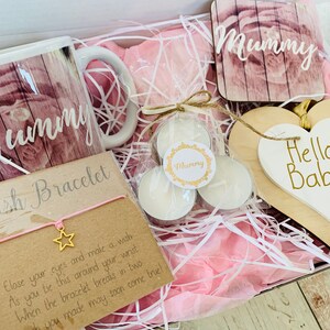 May include: A gift box filled with pink tissue paper and various items, including a mug with the word "Mummy" on it, a small wooden sign with the word "Mummy" on it, a small wooden sign with the words "Hello Baby" on it, a small card with the words "Wish Bracelet" on it, and three small white tea lights.