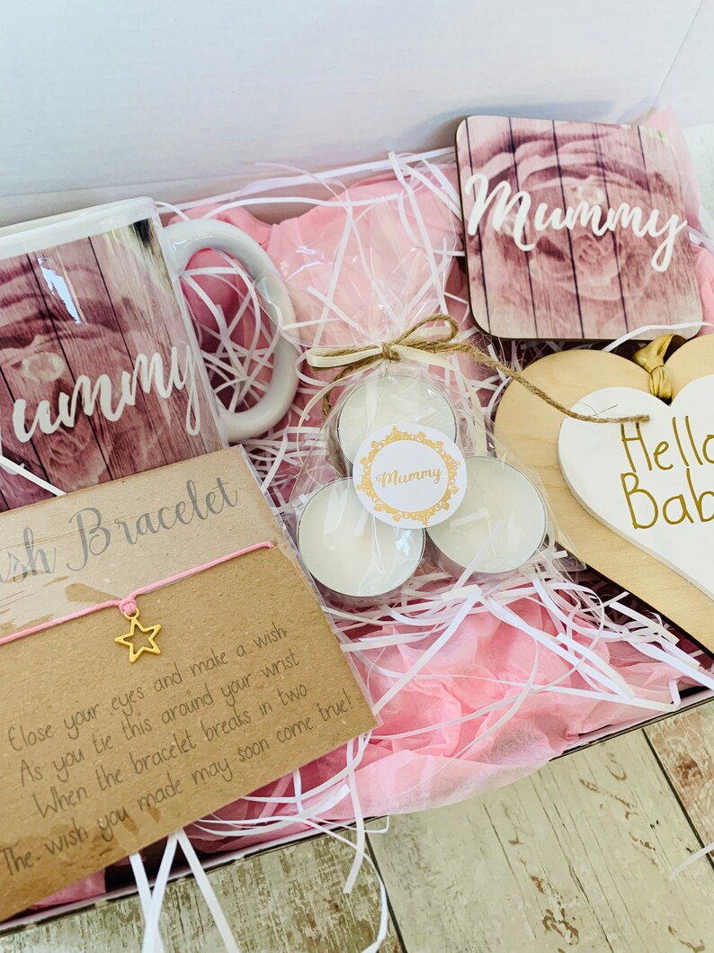 May include: A gift box filled with pink tissue paper and various items, including a mug with the word "Mummy" on it, a small wooden sign with the word "Mummy" on it, a small wooden sign with the words "Hello Baby" on it, a small card with the words "Wish Bracelet" on it, and three small white tea lights.