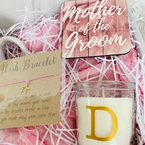 May include: A gift box filled with pink tissue paper, a white mug, a candle with a gold letter D, a wooden sign that says "Mother of the Groom", and a wish bracelet with a pink string and a gold star charm.
