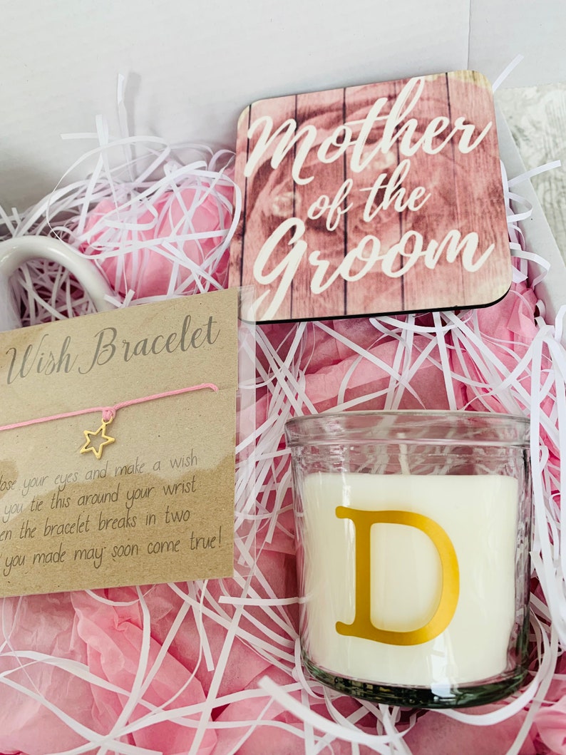 May include: A gift box filled with pink tissue paper, a white mug, a candle with a gold letter D, a wooden sign that says "Mother of the Groom", and a wish bracelet with a pink string and a gold star charm.