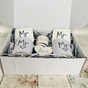 May include: A white gift box filled with white mugs, coasters, and a candle. The mugs have the word "Mr." printed on them, and the coasters have the word "Mr." printed on them. The candle has the words "&" and "Love" printed on it.