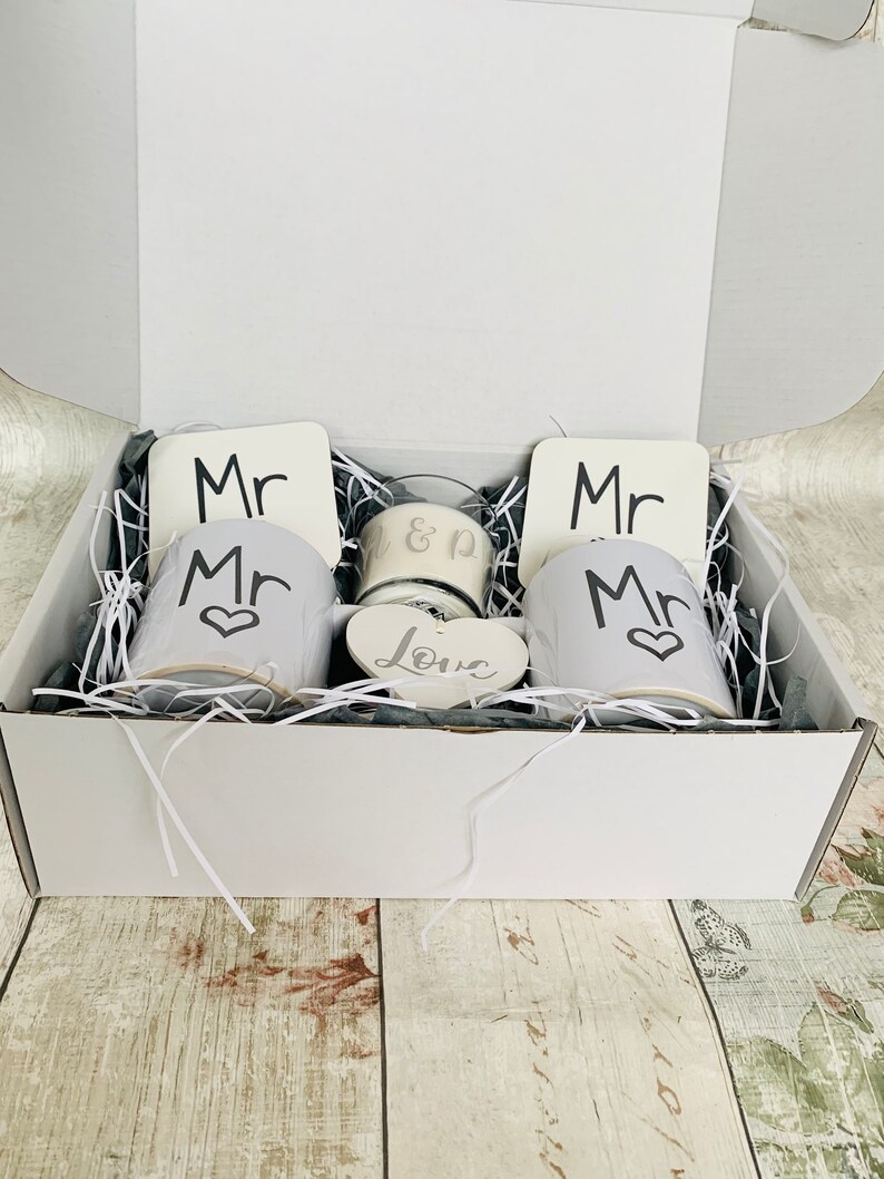 May include: A white gift box filled with white mugs, coasters, and a candle. The mugs have the word "Mr." printed on them, and the coasters have the word "Mr." printed on them. The candle has the words "&" and "Love" printed on it.