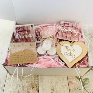 May include: A white gift box filled with pink tissue paper and a variety of gifts for a new mom. The gifts include a mug with the word "Mummy" on it, a wooden heart with the words "Hello Baby" on it, a wish bracelet, and two small candles.