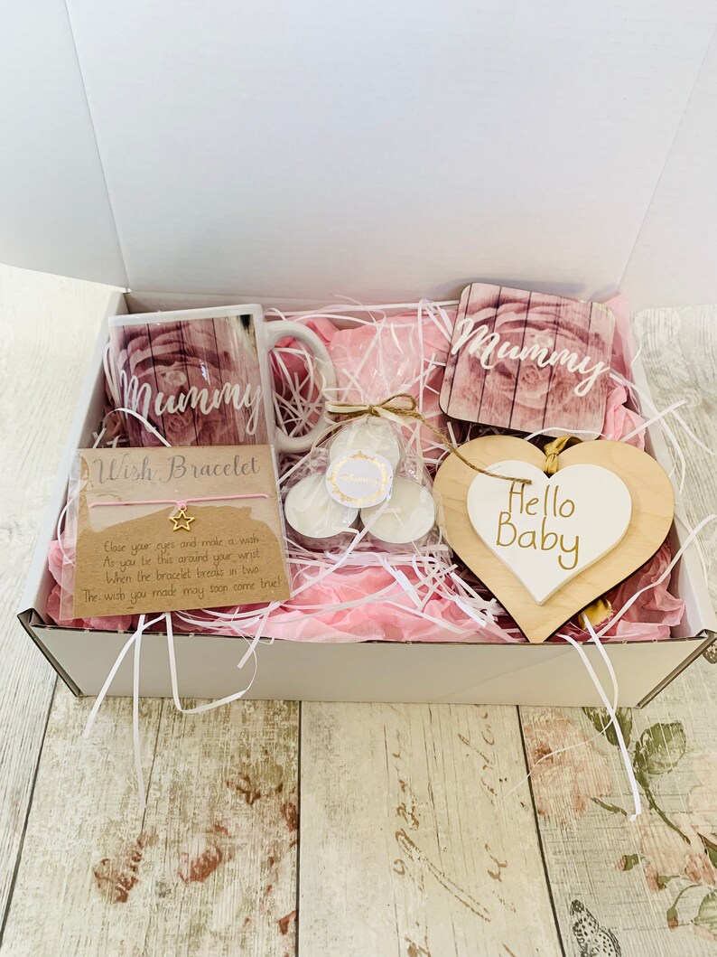 May include: A white gift box filled with pink tissue paper and a variety of gifts for a new mom. The gifts include a mug with the word "Mummy" on it, a wooden heart with the words "Hello Baby" on it, a wish bracelet, and two small candles.