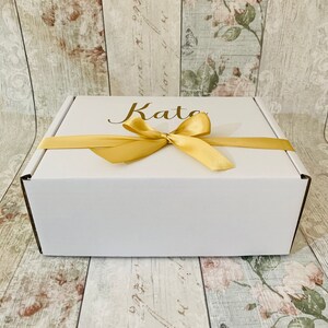 May include: White gift box with a gold ribbon and the name "Kate" written on the top.