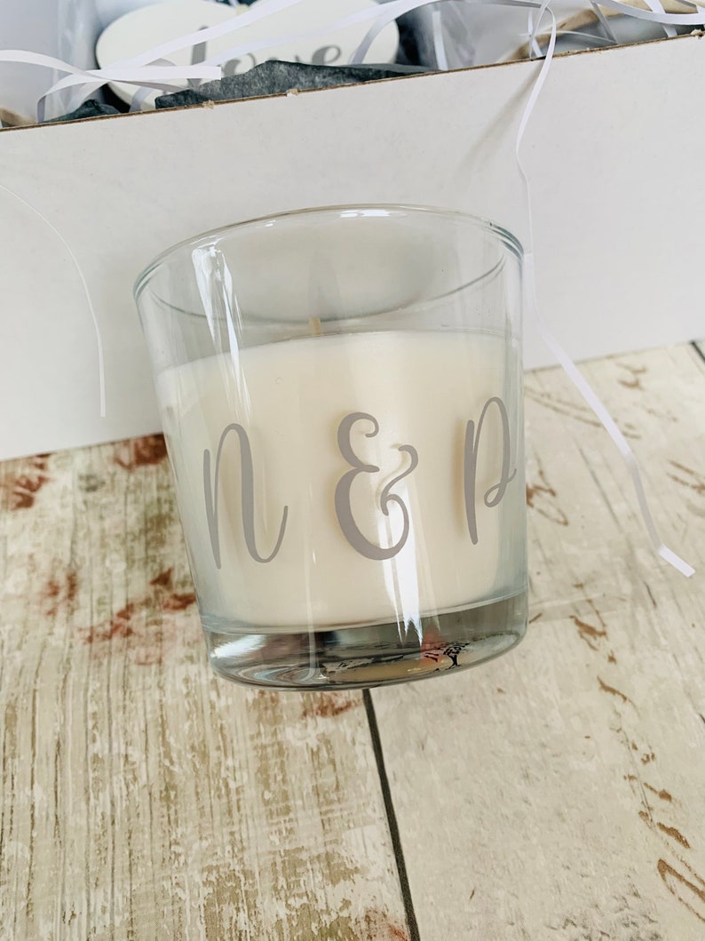 May include: A clear glass candle jar with a white candle inside. The candle has the letters "N & D" printed on the side in gray.