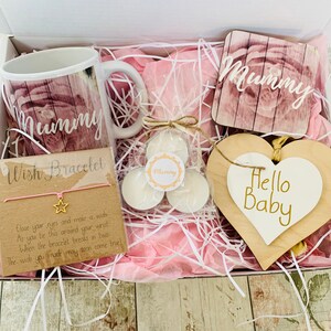 May include: A pink and white gift box filled with a pink and white mug, two small white candles, a pink and white coaster with the word "Mummy" on it, a pink wish bracelet, and a white heart-shaped wooden sign with the words "Hello Baby" on it.