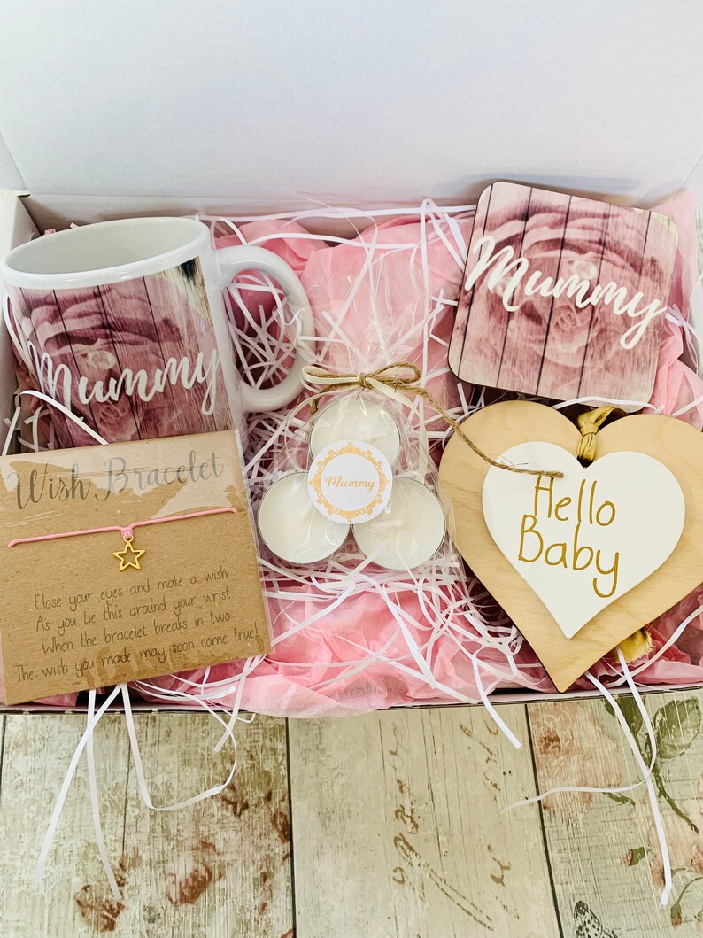 May include: A pink and white gift box filled with a pink and white mug, two small white candles, a pink and white coaster with the word "Mummy" on it, a pink wish bracelet, and a white heart-shaped wooden sign with the words "Hello Baby" on it.