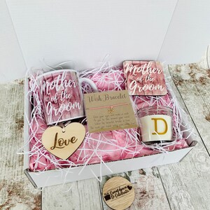 May include: A gift box filled with pink tissue paper and a variety of items for a mother of the groom. The items include a mug, a coaster, a candle, a wooden heart, and a wish bracelet. The mug and coaster feature the text "Mother of the Groom". The candle has the letter "D" on it. The wooden heart says "Love". The wish bracelet has a tag that says "Close your eyes and make a wish as you tie this around your wrist. When the bracelet breaks in two the wish you made may soon come true."