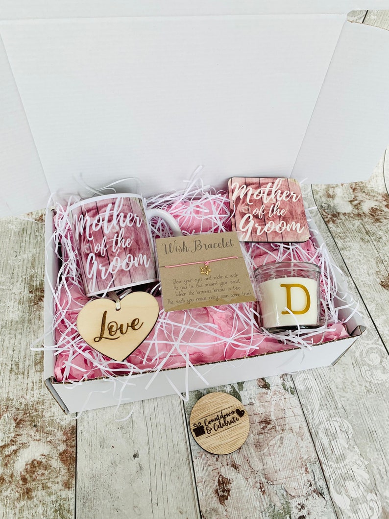 May include: A gift box filled with pink tissue paper and a variety of items for a mother of the groom. The items include a mug, a coaster, a candle, a wooden heart, and a wish bracelet. The mug and coaster feature the text "Mother of the Groom". The candle has the letter "D" on it. The wooden heart says "Love". The wish bracelet has a tag that says "Close your eyes and make a wish as you tie this around your wrist. When the bracelet breaks in two the wish you made may soon come true."