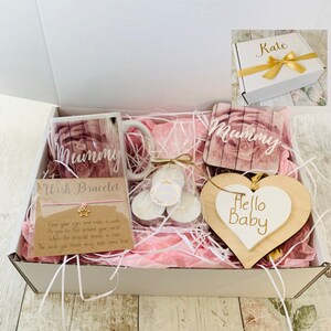 May include: A white gift box filled with pink tissue paper and various gifts. The gifts include a mug with the word "Mummy" on it, a wooden heart with the words "Hello Baby" on it, a coaster with the word "Mummy" on it, and a wish bracelet. The box is labeled with the name "Kate".