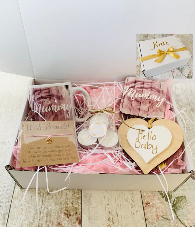 May include: A white gift box filled with pink tissue paper and various gifts. The gifts include a mug with the word "Mummy" on it, a wooden heart with the words "Hello Baby" on it, a coaster with the word "Mummy" on it, and a wish bracelet. The box is labeled with the name "Kate".