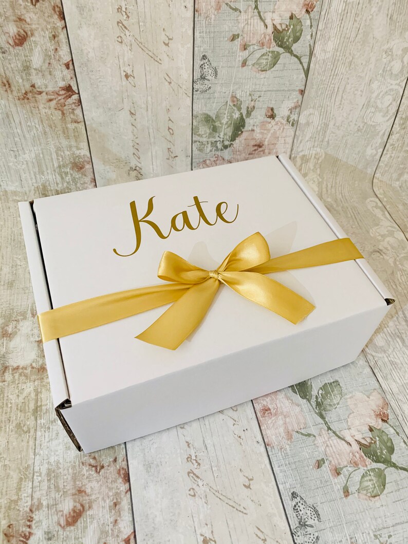 May include: A white gift box with a gold ribbon and bow. The box is personalized with the name "Kate" in gold lettering.