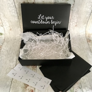 May include: A black gift box with a white inscription that reads "Let your countdown begin!" The box is open and filled with white shredded paper. There are ten numbered tags, each with the text "days to go" written on them, placed outside the box.