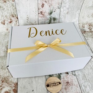 May include: A white gift box with a gold ribbon and the name "Denice" printed on the top. A small wooden circle with the text "Countdown & Celebrate" is in the foreground.