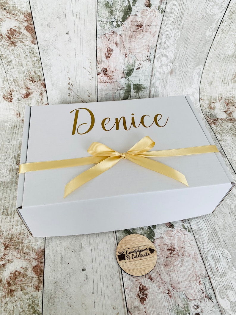May include: A white gift box with a gold ribbon and the name "Denice" printed on the top. A small wooden circle with the text "Countdown & Celebrate" is in the foreground.