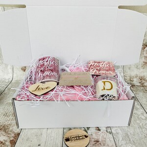May include: A white gift box filled with pink tissue paper and wedding-themed gifts. The gifts include two mugs with the text "Mother of the Groom", a wooden heart with the word "Love", a candle with the letter "D", and a wooden circle with the text "Countdown & Celebrate".