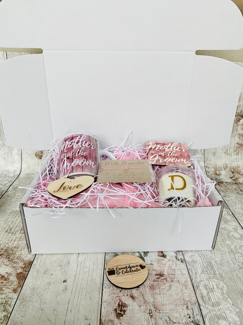 May include: A white gift box filled with pink tissue paper and wedding-themed gifts. The gifts include two mugs with the text "Mother of the Groom", a wooden heart with the word "Love", a candle with the letter "D", and a wooden circle with the text "Countdown & Celebrate".