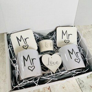 May include: A white gift box filled with a white gift basket containing two white mugs with the word "Mr" and a heart, two white coasters with the word "Mr" and a heart, a white heart with the word "Love", and a glass candle with the words "Mr & Mr".