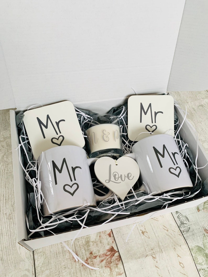 May include: A white gift box filled with a white gift basket containing two white mugs with the word "Mr" and a heart, two white coasters with the word "Mr" and a heart, a white heart with the word "Love", and a glass candle with the words "Mr & Mr".