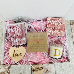May include: A gift box filled with pink tissue paper and a variety of items, including a mug that says "Mother of the Groom", a wooden heart that says "Love", a wish bracelet, a candle with the letter "D" on it, and a wooden coaster that says "Mother of the Groom".