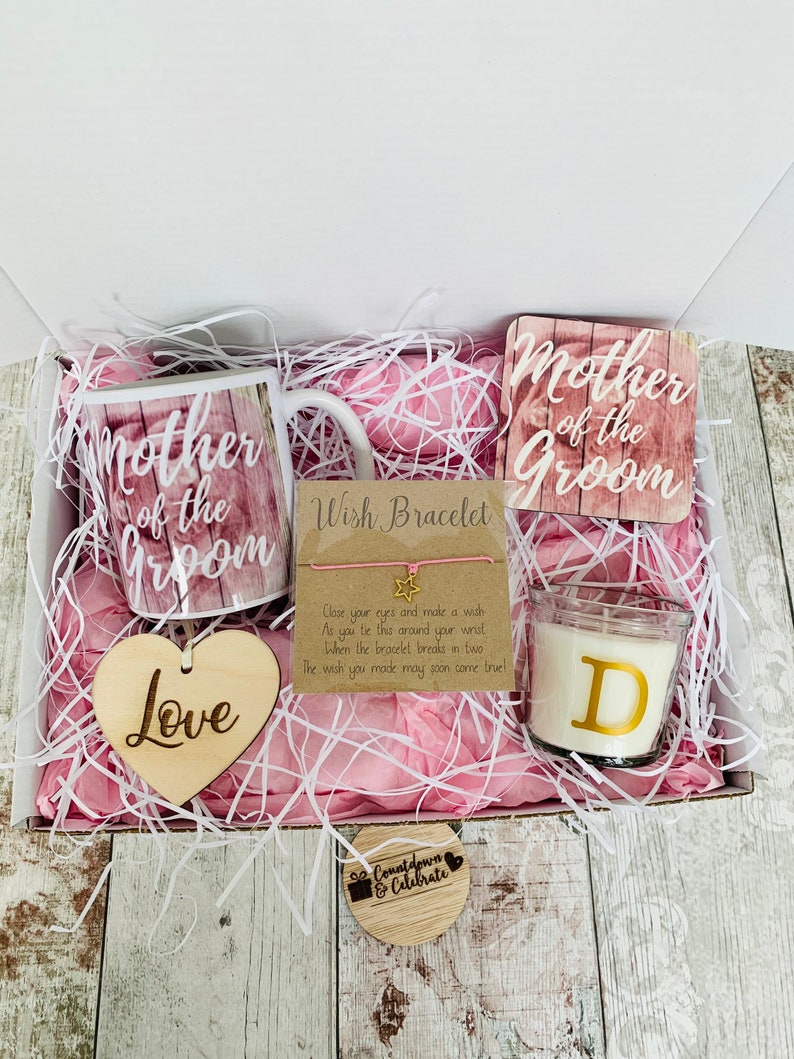 May include: A gift box filled with pink tissue paper and a variety of items, including a mug that says "Mother of the Groom", a wooden heart that says "Love", a wish bracelet, a candle with the letter "D" on it, and a wooden coaster that says "Mother of the Groom".