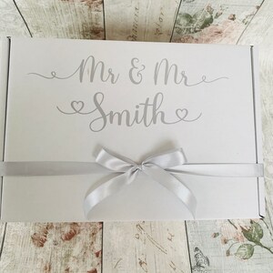 May include: White gift box with a silver ribbon and bow. The box is decorated with the text "Mr & Mr Smith" in a silver script font.