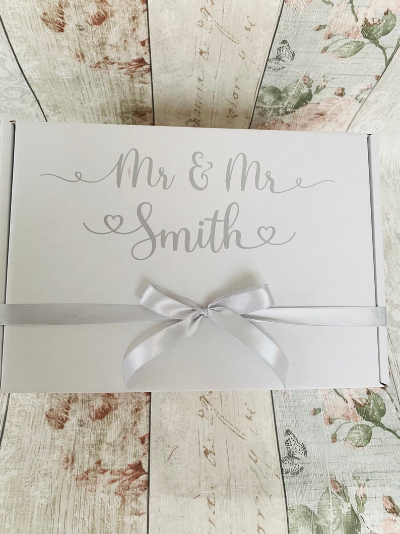May include: White gift box with a silver ribbon and bow. The box is decorated with the text "Mr & Mr Smith" in a silver script font.
