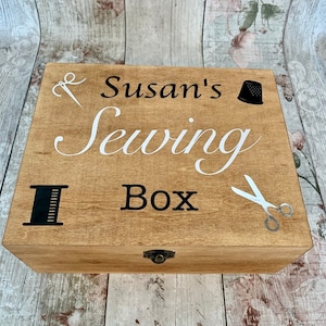 May include: A wooden sewing box with a hinged lid and a brass latch. The box is decorated with the text "Susan's Sewing Box" in white lettering and black and white illustrations of sewing tools.