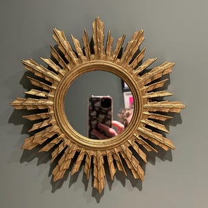 K&ouml;nnte beinhalten: Ein goldener Sonnenspiegel mit einem runden Spiegel in der Mitte. Der Spiegel ist von einem dekorativen Rahmen mit einem Sonnenstrahlendesign umgeben.