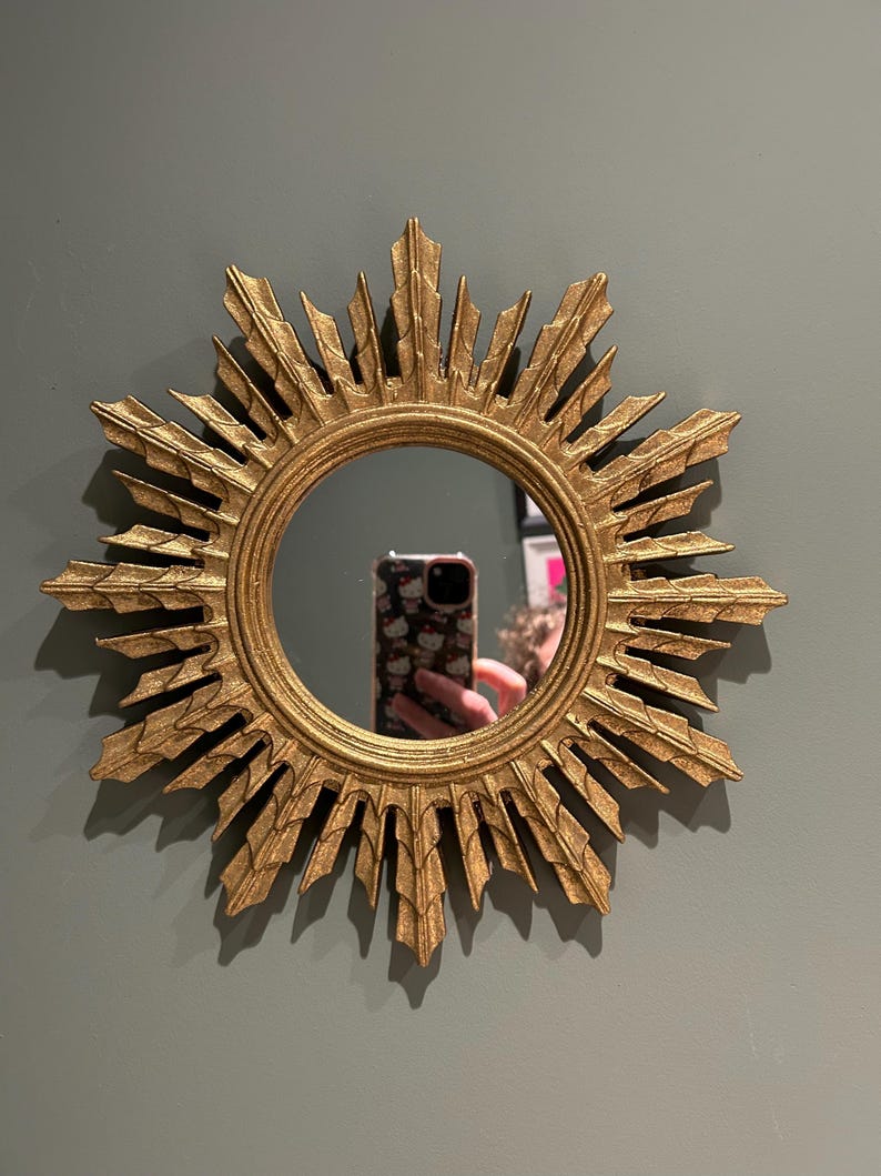 K&ouml;nnte beinhalten: Ein goldener Sonnenspiegel mit einem runden Spiegel in der Mitte. Der Spiegel ist von einem dekorativen Rahmen mit einem Sonnenstrahlendesign umgeben.
