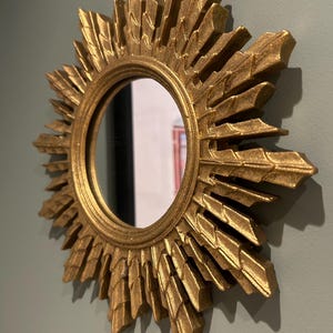 K&ouml;nnte beinhalten: Ein goldener Sonnenspiegel mit einer runden, reflektierenden Oberfl&auml;che. Der Spiegel hat einen dekorativen, verzierten Rahmen mit einem Sonnenstrahlendesign.