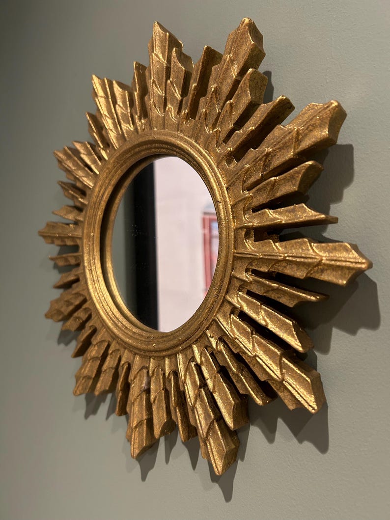 K&ouml;nnte beinhalten: Ein goldener Sonnenspiegel mit einer runden, reflektierenden Oberfl&auml;che. Der Spiegel hat einen dekorativen, verzierten Rahmen mit einem Sonnenstrahlendesign.