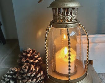 May include: A gold-colored lantern with a lit candle inside, featuring a clear glass enclosure and decorative metalwork. The lantern is accompanied by several pine cones, creating a warm, inviting ambiance. The lantern has a handle for carrying.