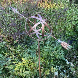 May include: A metal garden sculpture in the shape of an arrow pointing to a sphere. The sphere is made of metal and has a geometric design. The sculpture is brown and is set against a background of green foliage.