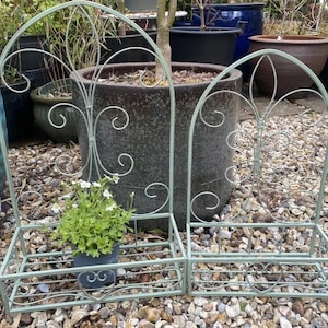 May include: Two decorative metal wall planters with a light green finish and ornate scrollwork. The planters have a shelf for plants and are mounted on a wall. The planters are filled with small white flowers and green plants.