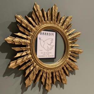 K&ouml;nnte beinhalten: Ein goldener Sonnenspiegel mit einem runden Spiegel in der Mitte. Der Spiegel reflektiert eine Schwarzwei&szlig;zeichnung einer Taube mit dem Wort "PICASSO" dar&uuml;ber.