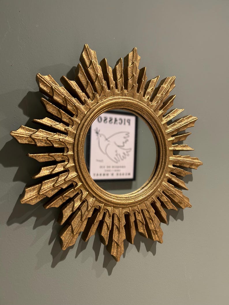 K&ouml;nnte beinhalten: Ein goldener Sonnenspiegel mit einem runden Spiegel in der Mitte. Der Spiegel reflektiert eine Schwarzwei&szlig;zeichnung einer Taube mit dem Wort "PICASSO" dar&uuml;ber.