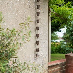 May include: A decorative rain chain featuring small, dark metal buckets linked together. The chain is suspended against a textured concrete wall, with foliage in the foreground and background. A bell is at the base.