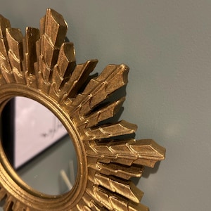 K&ouml;nnte beinhalten: Goldener Sonnenstrahlenspiegel mit einer runden, reflektierenden Oberfl&auml;che. Der Spiegel hat einen dekorativen, verzierten Rahmen mit einem Sternburst-Design.