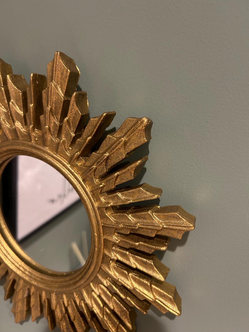 K&ouml;nnte beinhalten: Goldener Sonnenstrahlenspiegel mit einer runden, reflektierenden Oberfl&auml;che. Der Spiegel hat einen dekorativen, verzierten Rahmen mit einem Sternburst-Design.