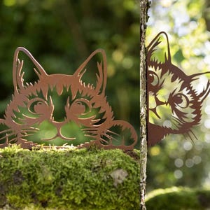 May include: Two metal cat silhouettes, one peeking from behind a tree trunk and the other looking over a mossy rock. Both cats are rusty brown in colour and have a whimsical, cartoonish style.