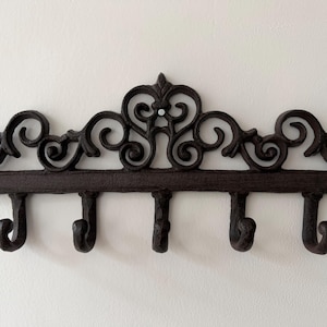 May include: Ornate, dark brown metal wall hook rack with five hooks. The rack features a decorative, scrolled design across the top. Perfect for entryways, bedrooms, or bathrooms. Measures approximately 30 cm wide.