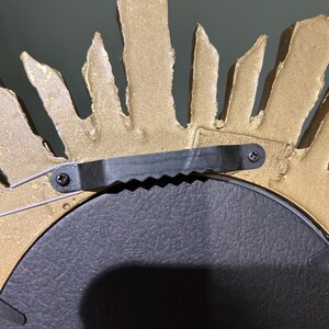 K&ouml;nnte beinhalten: Goldfarbener Sonnenstrahlenspiegel mit schwarzem Metallrahmen und Aufh&auml;ngevorrichtung. Der Spiegel hat eine strukturierte, raue Oberfl&auml;che.