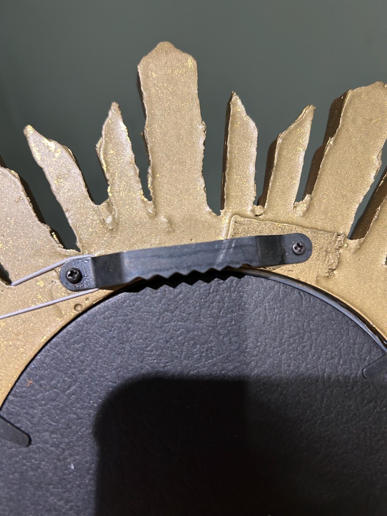 K&ouml;nnte beinhalten: Goldfarbener Sonnenstrahlenspiegel mit schwarzem Metallrahmen und Aufh&auml;ngevorrichtung. Der Spiegel hat eine strukturierte, raue Oberfl&auml;che.