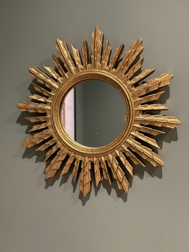 K&ouml;nnte beinhalten: Ein goldener Sonnenspiegel mit einer runden, reflektierenden Oberfl&auml;che. Der Spiegel hat einen dekorativen, verzierten Rahmen mit einem Sonnenstrahlendesign.
